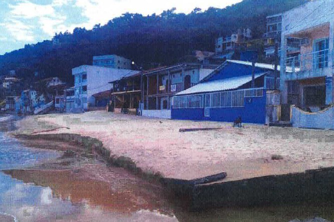 A imagem mostra uma praia com várias construções ao longo da orla. Os edifícios variam em tamanho e cor, alguns são de vários andares e outros térreos. A praia tem sinais de erosão próximos à água. Ao fundo, há uma colina ou montanha com mais construções visíveis.