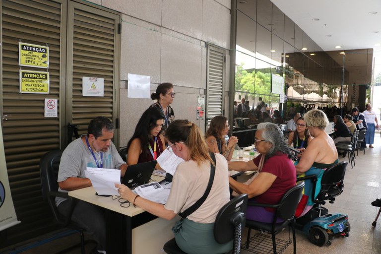 A imagem mostra um grupo de pessoas sentadas em mesas em uma área externa coberta. No primeiro plano, à esquerda, há um homem e uma mulher sentados em frente a um laptop, examinando papéis. Outra mulher está sentada à frente deles, de costas, lendo documentos. Mais à direita, duas mulheres, uma delas mais velha com cabelos grisalhos, estão sentadas e conversando. Atrás das mesas, uma mulher em pé supervisiona a área.
