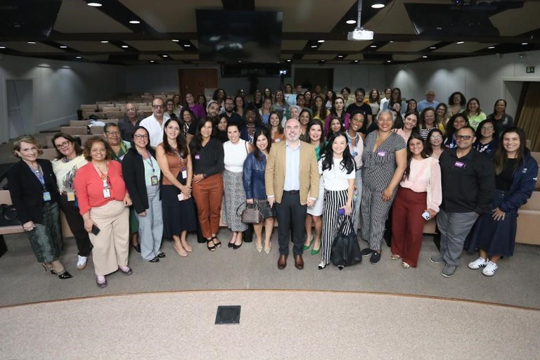 A imagem mostra um grande grupo de pessoas, aparentemente profissionais de diversas áreas, posando para uma foto em um auditório. A maioria das pessoas está em pé, formando um semicírculo em frente a uma mesa de madeira. No centro da foto, em primeiro plano, um homem de camisa social clara e paletó de cor cáqui se destaca, cercado por mulheres com diferentes vestimentas casuais e sociais. Ao fundo, é possível ver as cadeiras vazias do auditório e telas de projeção. A foto transmite a impressão de um evento profissional, como um fórum ou encontro.