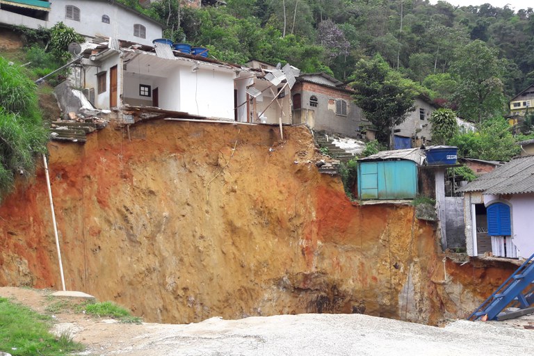 Fotografia externa que mostra um grave deslizamento de terra em uma área residencial na encosta de um morro. Uma enorme cratera de terra avermelhada e amarelada expõe um grande barranco vertical após o colapso do solo. Na parte superior da falésia, casas foram parcialmente destruídas e ficaram à beira do precipício; uma casa branca no centro teve sua parede frontal e assoalho arrancados, revelando o interior dos cômodos e partes do telhado de chapa metálica retorcido. Ao lado, outras casas simples permanecem erguidas perto do abismo, incluindo uma pequena construção de metal azul. Ao fundo, a vegetação verde do morro se estende até o topo sob um céu claro.