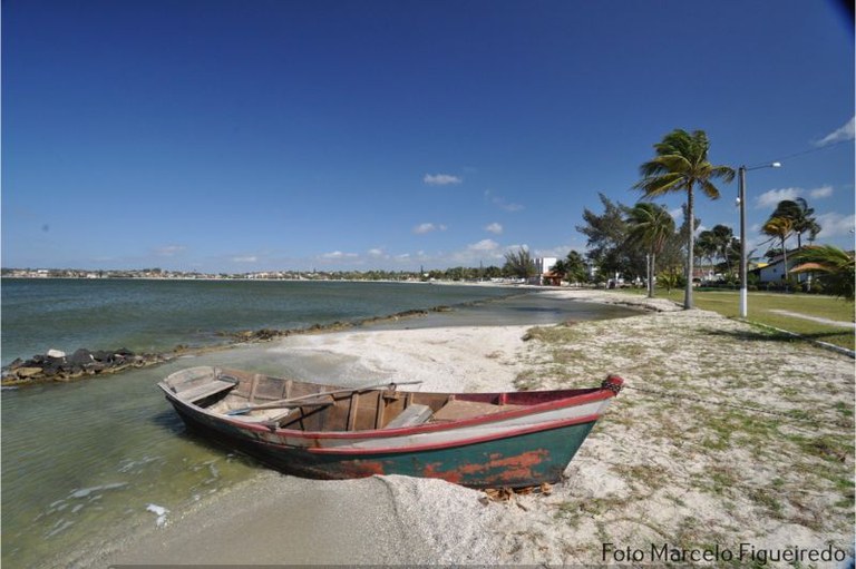 Foto da orla da praia mostra o mar, um barco de pescador pequeno na areia, coqueiros e casas ao fundo
