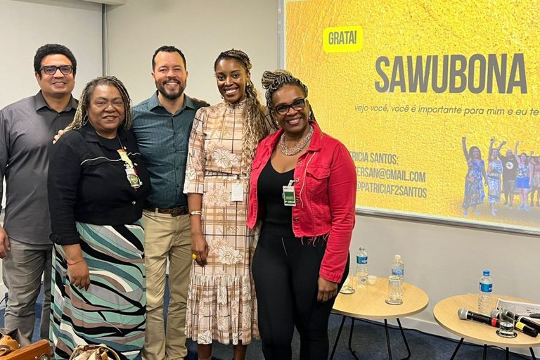 Cinco pessoas estão lado a lado, posando sorridentes para a foto em uma sala de palestras. Da esquerda para a direita, há três mulheres e dois homens. Todos usam crachás funcionais presos à roupa. Ao fundo, na parede, há uma tela de projeção com o título “SAWUBONA” em letras grandes, seguido da frase: “vejo você, você é importante para mim e eu te valorizo”. No canto superior esquerdo da tela, há um balão amarelo com a palavra “GRATA!”. Abaixo do texto, estão os contatos da palestrante Patricia Santos. À frente da tela, há duas mesinhas com garrafas de água, microfones e papéis, sugerindo o encerramento de uma roda de conversa ou palestra. O ambiente é iluminado e transmite um clima acolhedor e de celebração.