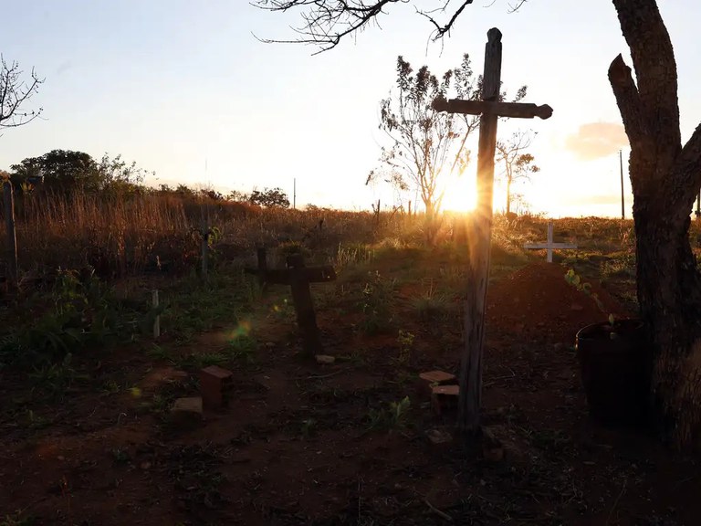 A imagem mostra uma paisagem de cemitério com uma cruz de madeira rústica e alta fincada no chão. A luz do sol brilha intensamente por trás dela, criando um forte efeito de raios de sol e um contraluz. O solo é de terra batida
