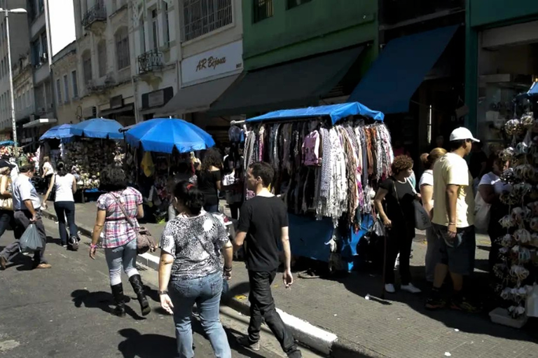 Fotografia em ângulo plongée e plano geral que mostra uma movimentada rua de comércio popular a céu aberto com pedestres caminhando pelo asfalto e pelas calçadas. Em primeiro plano, pessoas de costas andam em direção ao fundo da via, vestindo roupas casuais. Ao lado direito e ao longo da rua, veem-se barracas de camelôs cobertas por lonas e guarda-sóis azuis, exibindo mercadorias como lenços, roupas e bijuterias penduradas. Ao fundo, prédios comerciais urbanos com marquises e toldos emolduram o cenário ensolarado, projetando sombras marcadas no chão.