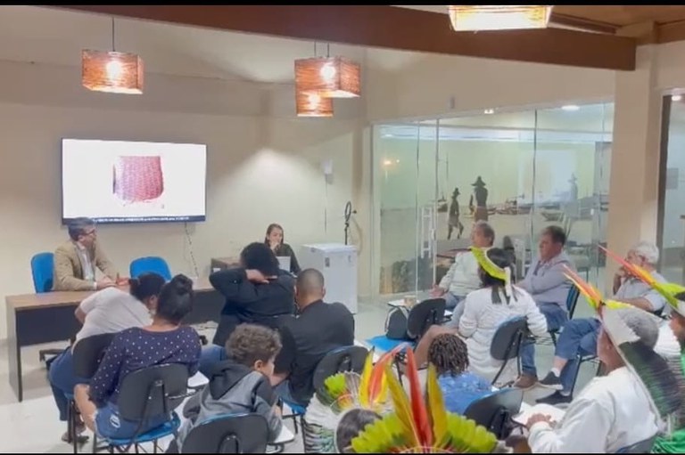 A imagem anexa mostra uma reunião ou apresentação em ambiente interno com aproximadamente 15 a 20 pessoas. O foco da sala parece estar em uma tela de televisão/monitor à esquerda, que exibe uma imagem em close-up de um objeto com textura de cestaria ou tecido. À frente da tela, um homem de blazer e óculos está sentado a uma mesa, conversando com uma mulher que está de pé ou sentada na mesma mesa. A maioria dos participantes está sentada em cadeiras dispostas em filas, e alguns deles, em primeiro plano à direita, usam adornos de cabeça indígenas com penas coloridas, indicando a participação de membros de povos originários no evento. O ambiente é bem iluminado, tem um aspecto moderno e, ao fundo, há uma parede de vidro que permite ver uma área externa ou um diorama com figuras que parecem representar pessoas.