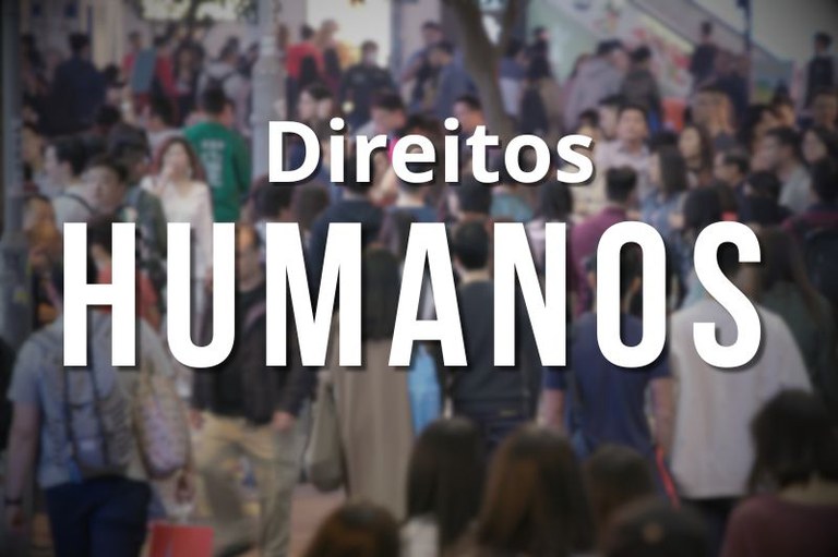 Texto "Direitos HUMANOS" em letras brancas em destaque, sobreposto a uma foto intencionalmente desfocada de uma grande multidão de pessoas caminhando em um espaço público.