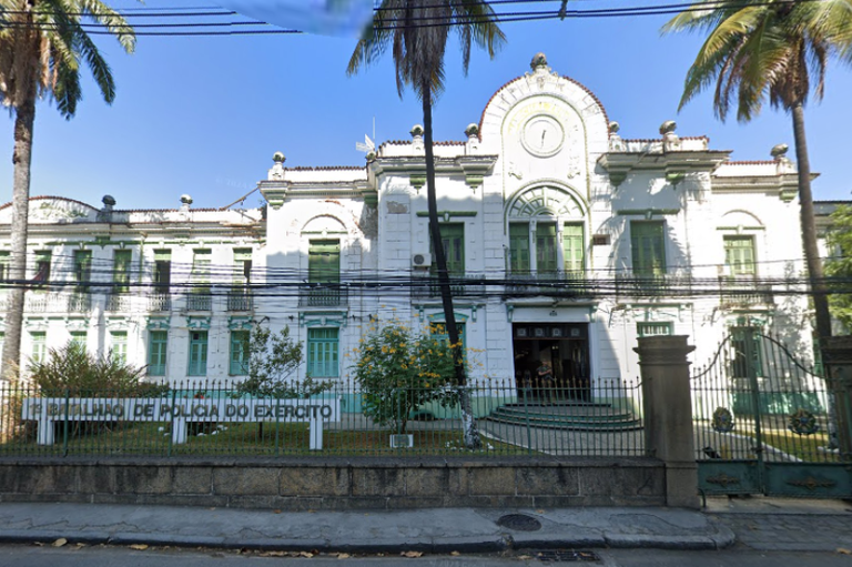 Foto da fachada do prédio do 1º Batalhão de Polícia do Exército; prédio tem estilo de arquitetura antigo, com fachada em branco e verde, cercado por muro baixo e grades.