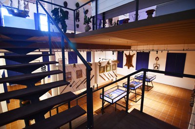 Fotografia em perspectiva superior do interior de um museu, capturada a partir do patamar de uma escada com estrutura metálica e degraus escuros. O salão tem piso de cerâmica tom tijolo e paredes brancas com portas e janelas azuis ao fundo. No térreo, expostos no centro e nas paredes, há painéis brancos com quadros, arcos, flechas, ferramentas indígenas, uma pele de felino pendurada e vitrines horizontais de vidro com bordas azuis contendo pequenos artefatos arqueológicos. O mezanino de teto de madeira abriga vasos de plantas, uma réplica de embarcação antiga e objetos étnicos dispostos nas paredes superiores.