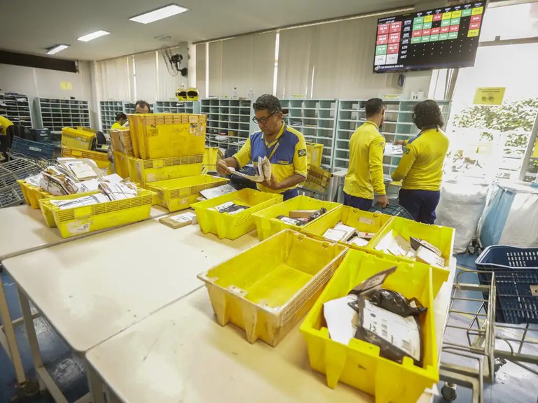 A imagem mostra funcionários dos Correios trabalhando na triagem de encomendas dentro de um centro operacional, utilizando caixas plásticas amarelas para organizar as cartas e pacotes.