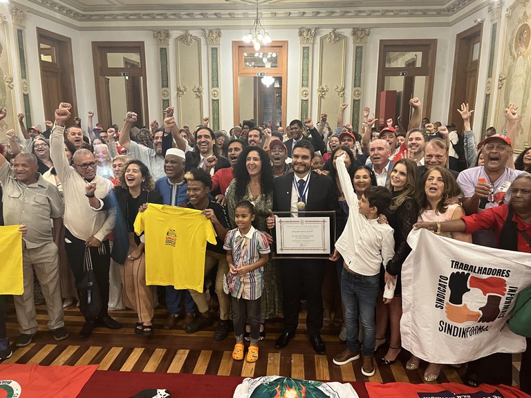 A imagem mostra uma grande cerimônia em um salão elegante, com paredes ornamentadas e lustres no teto. No centro, o procurador Julio Araujo, usando terno, segura um diploma e uma medalha, cercado por muitas pessoas que levantam os braços em gesto de comemoração. Há diversidade de idades, gêneros e etnias entre os presentes. Algumas pessoas seguram camisetas e bandeiras de movimentos sindicais e sociais.