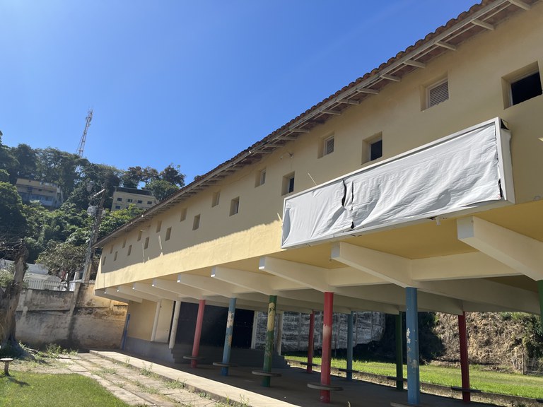 Fotografia em plano aberto de um edifício escolar de dois pavimentos, visto em perspectiva lateral. A construção possui fachada em tom amarelo-claro, telhado de cerâmica e diversas pequenas janelas quadradas distribuídas ao longo do andar superior. Na parte inferior, o prédio é sustentado por pilares pintados em cores variadas — azul, vermelho, verde e amarelo — formando um espaço coberto e aberto. À direita da imagem, há uma grande estrutura retangular branca, semelhante a um letreiro ou painel, com a lona rasgada no centro. Em primeiro plano, aparecem uma área gramada e um caminho de concreto com vegetação crescendo entre as placas. Ao fundo, à esquerda, há um morro com árvores, postes, uma torre de telecomunicações e edificações. O céu está completamente azul, sem nuvens, e a iluminação indica um dia ensolarado.