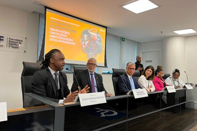 Fotografia em ambiente interno mostra seis pessoas sentadas lado a lado em uma mesa retangular com placas de identificação à frente. Ao fundo, no centro, há uma tela de projeção exibindo cartaz amarelo sobre seminário contra o racismo, com a logo do MPF ao lado esquerdo e bandeiras do Brasil e de estados à esquerda. O grupo é composto por três homens de terno e três mulheres; a mulher mais à direita usa blazer rosa, óculos de armação branca e colar largo.