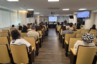 Foto em ambiente interno de um auditório mostra cadeiras com o público e ao fundo um telão com uma mesa na frente