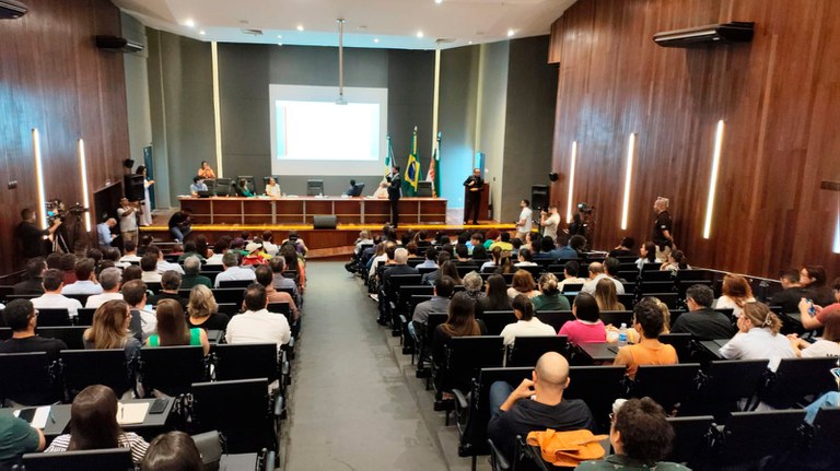 Imagem do auditório, em direção ao palco, vendo-se na parte de baixo o público sentado, de costas, e sobre o palco o procurador da República Camões Boaventura fazendo uma apresentação ao microfone, utilizando um telão por trás da mesa principal