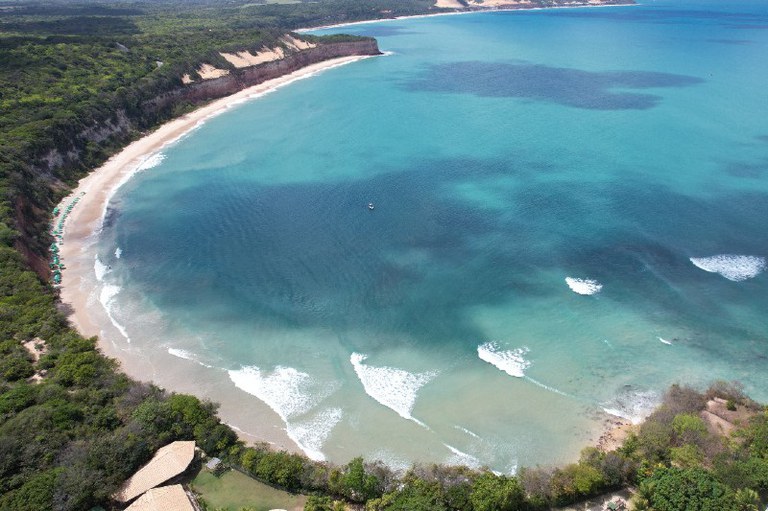 Foto das falésias de Pipa, tomada de cima, de um drone, a partir do continente, de onde se vê a vegetação por sobre as falésias, bem como parte da praia e o mar em frente, formando uma curva côncava na praia"