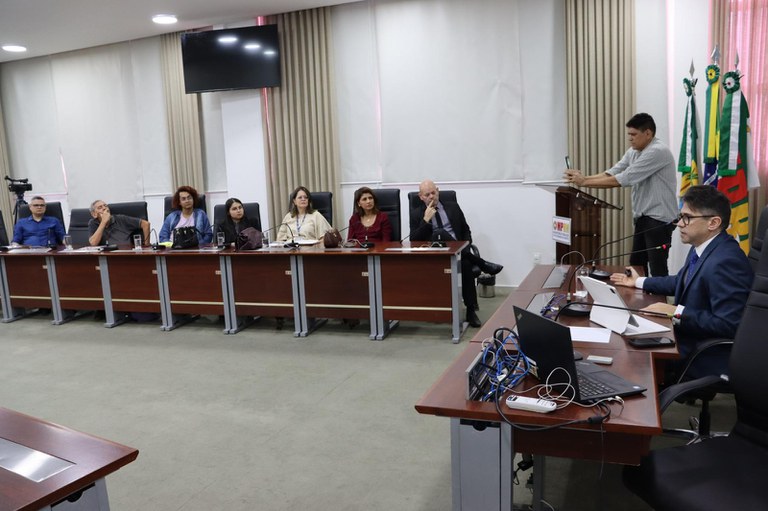 No auditório do MPRN, o procurador regional eleitoral Fernando Rocha fala ao microfone, de terno, sentado diante de uma mesa com notebook, tablet, papéis e outros equipamentos. À esquerda, sete pessoas acompanham a apresentação, sentadas atrás de uma bancada. Ao fundo, outro homem registra o momento com um celular próximo a um púlpito. O ambiente possui cortinas claras, uma televisão desligada e bandeiras institucionais.