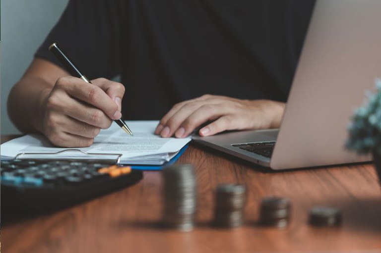 Foto em detalhe de uma pessoa conferindo um documento com uma caneta, ao lado um notebook aberto, à frente uma calculadora e quatro pilhas de moedas