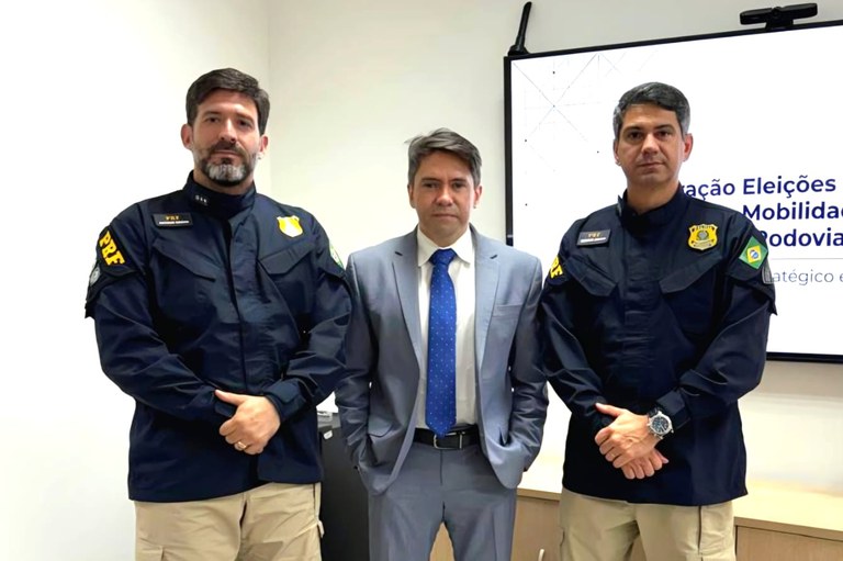 Três homens estão lado a lado, em pé, posicionados em frente a uma parede branca e ao lado de uma tela de apresentação. Ao centro, um homem veste terno cinza, camisa social clara e gravata azul com pequenas estampas; ele está com as mãos nos bolsos da calça. Ladeando-o, dois policiais rodoviários federais vestem o uniforme oficial da corporação, composto por camisa operacional preta de mangas compridas, com o distintivo do órgão no peito e a sigla "PRF" na manga, e calça bege. Ambos permanecem eretos com as mãos cruzadas à frente do corpo. Ao fundo, no canto superior direito, vê-se parte de uma tela digital exibindo a frase "Ação Eleições" e outros textos.. Reunião de alinhamento entre o procurador regional eleitoral, Fernando Rocha (centro), e o superintendente substituto da PRF no RN, Hudson Sérgio de Araújo (direita).