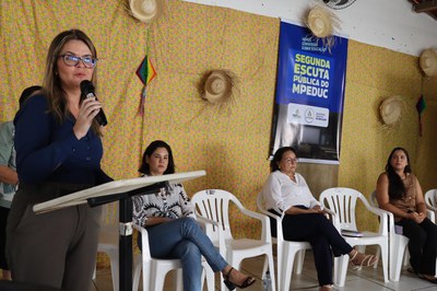 Fotografia colorida em plano médio-longo que registra um evento institucional. Em primeiro plano, à esquerda, uma mulher de cabelos castanhos claros e óculos de grau veste blusa de manga longa azul-marinho e calça social cinza, falando ao microfone em um púlpito branco. Ao fundo, três mulheres estão sentadas em cadeiras de plástico brancas alinhadas: a primeira veste camisa estampada em preto e branco e calça jeans; a segunda usa camisa branca e calça escura; e a terceira veste blusa marrom e calça preta. A parede atrás delas é decorada com um tecido estampado amarelo com pequenas flores coloridas, ornamentada com chapéus de palha e pequenos enfeites de festa junina, além de um banner vertical azul à direita com a inscrição "SEGUNDA ESCUTA PÚBLICA DO MPEDUC".