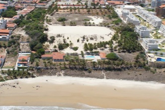 Foto aérea, tirada da praia para o litoral, mostra área da duna, com vegetação ao redor, e tendo em sua volta diversos imóveis, entre casas e condomínios. Abaixo na imagem aparece a linha da praia de Pirangi.