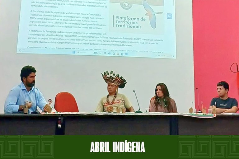 Imagem da mesa de diálogo do evento, sobre o palco de um auditório, com o procurador Camões Boaventura à esquerda, ao centro a juíza federal Gisele Leite e o cacique Carlos Potiguar e, à direita, o representante do DCE da UFRN, José Victor, todos sentados. À frente da mesa, um cesto e, por trás, um telão aparecendo imagens referente à plataforma Territórios Tradicionais.