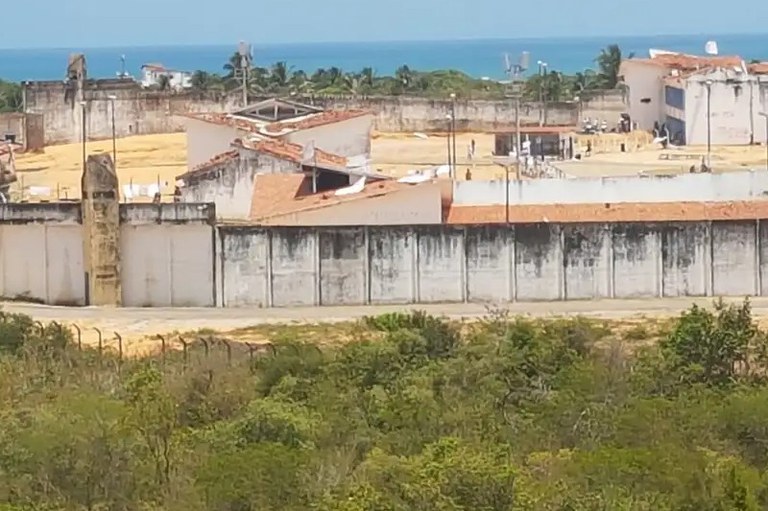 Foto da Penitenciária Estadual de Alcaçuz, tirada do lado externo, em lugar alto, em que se vê os muros da penitenciária, áreas abertas dentro do presídio e algumas construções