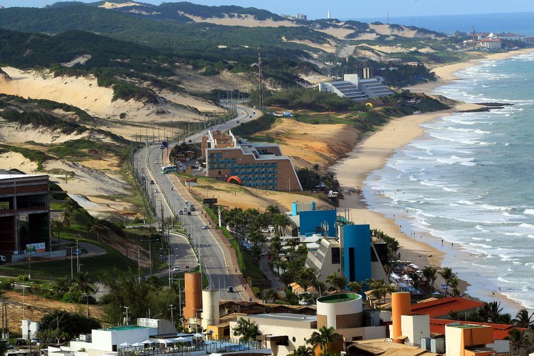 Imagem aérea, registrando parte da Via Costeira, no sentido Norte-Sul, aparecendo à esquerda as dunas e vegetação do Parque das Dunas; no centro a pista principal e hotéis; e à direita as praias e o mar. tudo do ponto de vista de quem se encontra acima do bairro de Ponta Negra.