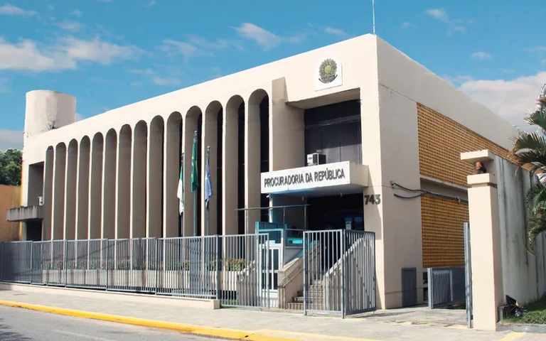 Imagem da fachada do prédio da PR/RN, bege com grades na frente, tendo acima o céu azul e observando-se parte da calçada em frente ao local
