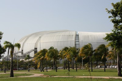 A imagem mostra uma grande estrutura de telhado branco e curvo, que se assemelha a uma concha, sob um céu claro. Em primeiro plano, há uma área gramada com fileiras de coqueiros e outras árvores, que parecem estar sendo balançadas pelo vento. Vários carros brancos estão estacionados ao fundo.