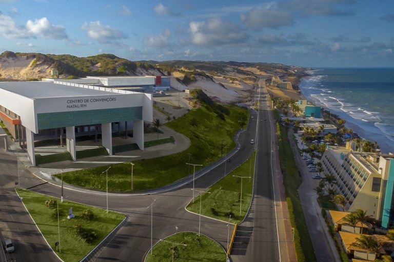Esta imagem aérea mostra uma bela vista panorâmica da Via Costeira em Natal, Rio Grande do Norte, destacando o moderno complexo do Centro de Convenções de Natal/RN à esquerda. O edifício possui uma fachada imponente branca com detalhes em verde e colunas estruturais elevadas sobre um gramado bem cuidado. A rodovia de duas pistas corta o centro da composição, estendendo-se em direção ao horizonte ladeada, do lado esquerdo, pelas famosas dunas de areia cobertas por vegetação nativa rasteira e, do lado direito, pela linha costeira com praias, hotéis, coqueiros e o mar azul com ondas suaves sob um céu parcialmente nublado.