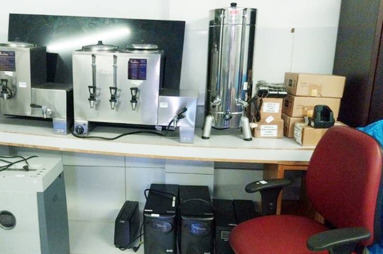 Foto mostra parte de um lote, com computadores e equipamentos em cima de uma mesa e uma cadeira ao lado da mesa