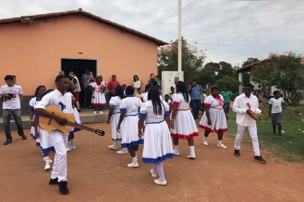 A imagem mostra um grupo de pessoas que está reunido ao ar livre. Algumas vestem trajes brancos com detalhes azuis e vermelhos e estão dançando. Há músicos tocando violão e tambor. Alguns espectadores assistem à apresentação.