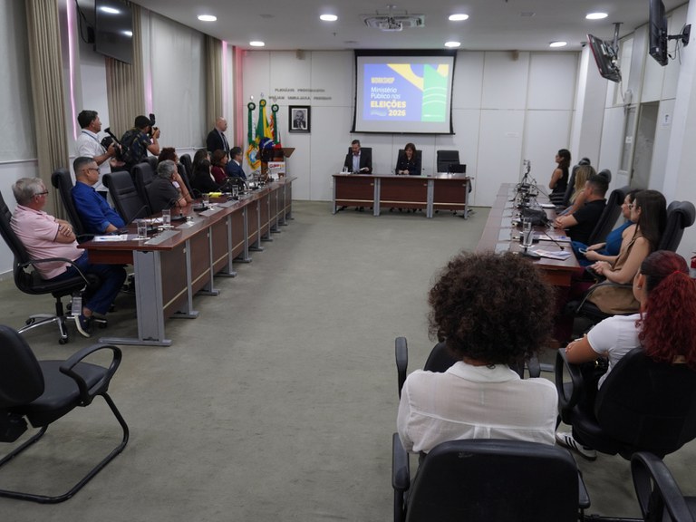 Auditório do MPRN, fotografado da entrada, tendo nas laterais pessoas sentadas em duas mesas, jornalistas e assessores, e tendo ao fundo a mesa principal, com a procuradora procuradora-geral de Justiça de adjunta (PGJA), Juliana Limeira, e o representando a Mídiacom, Tiago Lajus.