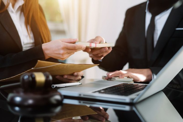 Imagem mostra um notebook na mesa, um homem e uma mulher e um maço de dinheiro