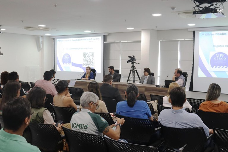 Fotografia de pessoas sentadas em cadeiras na platéia do auditório da sede do MPF em Natal (RN), com a mesa do evento ao fundo, com dois telões, em em cada lado. A mesa é composta, da esquerda pra direita, por Ilia Freire, Adriano Platiny, Gilberto Barroso e André Nunes.
