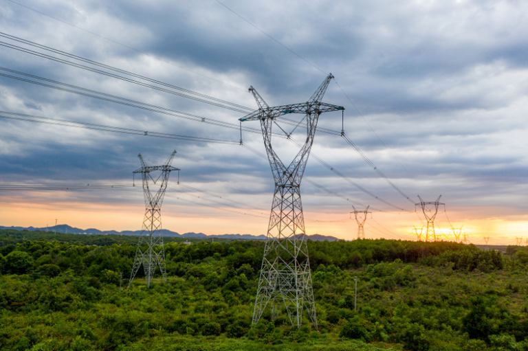 Fotografia de grandes torres metálicas de transmissão de energia elétrica que atravessam uma área de vegetação densa e verde. As torres sustentam diversas linhas de cabos sob um céu carregado de nuvens cinzentas, com o horizonte iluminado por um pôr do sol amarelado ao fundo.