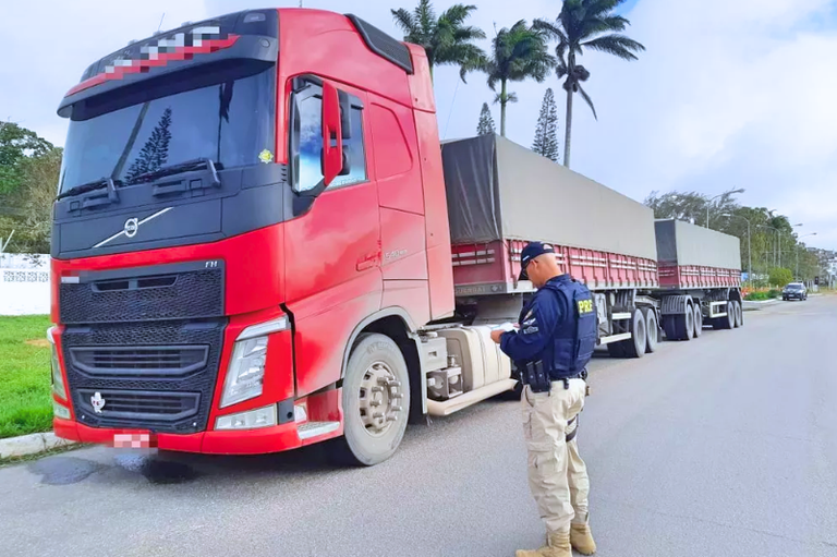 Caminhão-trator vermelho com semirreboques estacionado em uma via, enquanto um agente da Polícia Rodoviária Federal (PRF), uniformizado, realiza uma fiscalização ao lado do veículo.