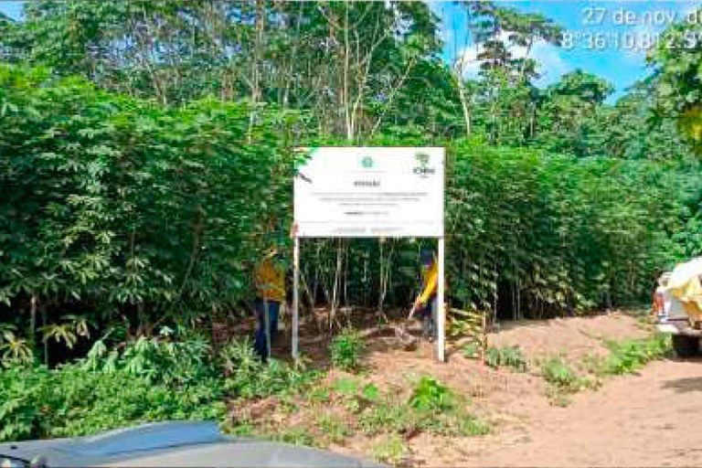 Foto mostra placa instalada em estrada aberta no meio de uma área verde, com bastante vegetação ao redor.