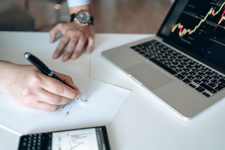 Foto mostra em detalhe uma pessoa escrevendo em um papel em uma mesa com um notebook e um celular
