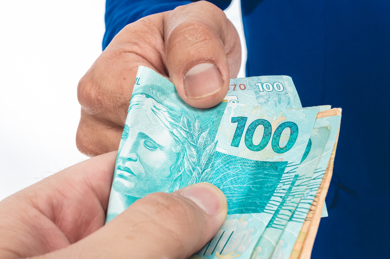 Foto mostra o detalhe da mão de uma pessoa vestindo terno azul e a mão de outra pessoa (que não aparece na foto) segurando um maço de dinheiro com notas de 100 e 50 reais