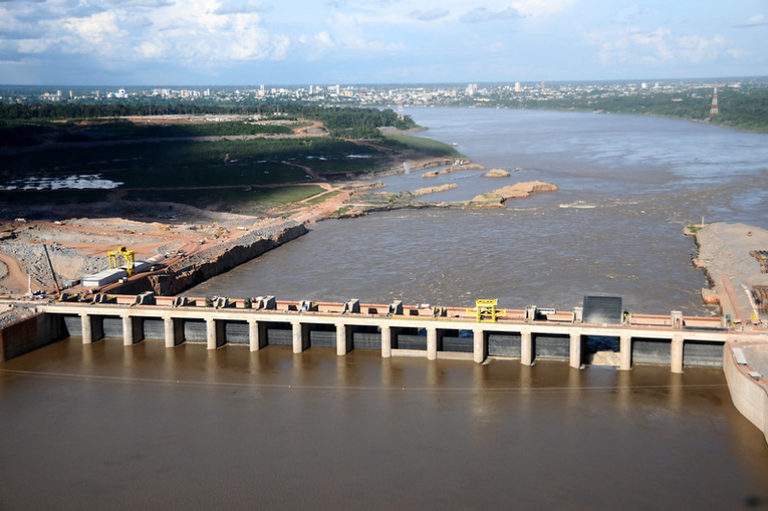 Vista aérea panorâmica da Usina Hidrelétrica de Santo Antônio instalada em um largo rio de águas barrentas, com a linha do horizonte revelando uma área urbana e vegetação sob um céu claro.