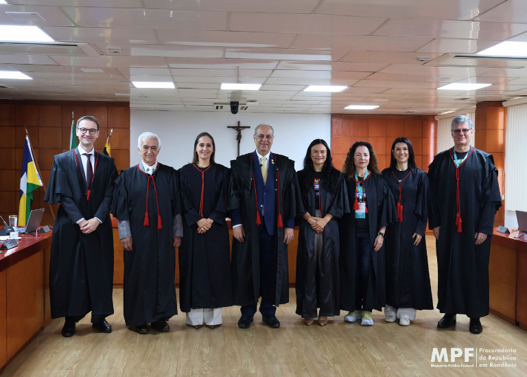 Fotografia de grupo com nove pessoas, entre homens e mulheres, usando vestes formais e togas pretas com detalhes vermelhos, em uma sala institucional com paredes de madeira, mesas e bandeiras ao fundo.
