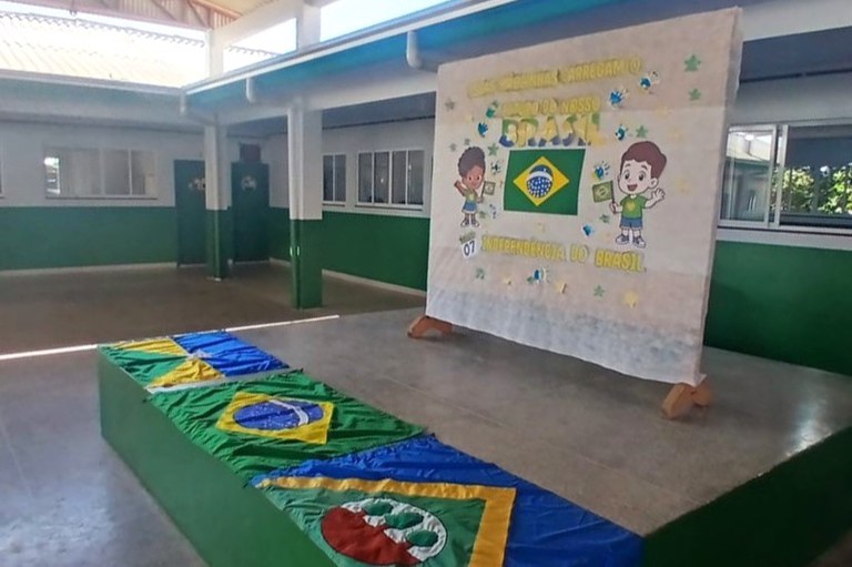 Fotografia colorida do pátio interno de uma escola, pintado nas cores verde e branco. A cena mostra uma área elevada que serve como palco com um banner temático. O banner, em tecido branco, exibe a Bandeira do Brasil centralizada e a frase "7 de Setembro: Dia da Independência do Brasil", ladeada por ilustrações de duas crianças sorrindo. À frente do palco, em uma estrutura retangular pintada de verde, estão estendidas e dobradas as bandeiras de dois estados brasileiros e a do Brasil, todas alinhadas e coloridas.
