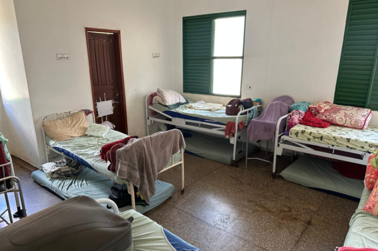 Quarto coletivo com várias camas de estilo hospitalar e colchões adicionais dispostos no chão sob os leitos. Ao fundo, veem-se janelas de ferro venezianas verdes e uma porta de madeira.