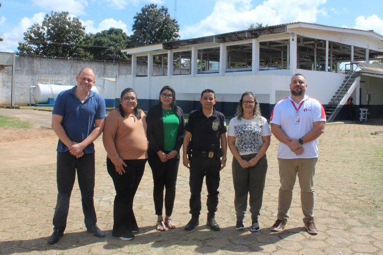 Fotografia do procurador da República Reginaldo Trindade, o diretor geral e mais quatro pessoas da equipe do Copen/RO, sendo três mulheres e um homem, todos em pé, dentro do pátio da unidade prisional, com o muro e um galpão ao fundo.