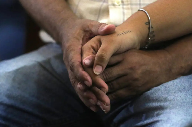 Fotografia em close de duas mãos entrelaçadas em um gesto de apoio e união. Uma das mãos possui tatuagens discretas no pulso e no dorso, repousando sobre as mãos de outra pessoa.