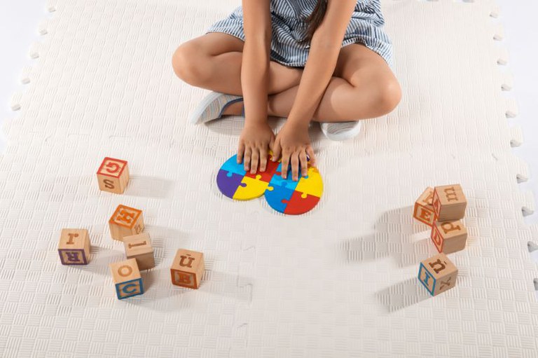 Foto mostra criança brincando no chão segundando um coração colorido com quebra-cabeças, símbolo do autismo.