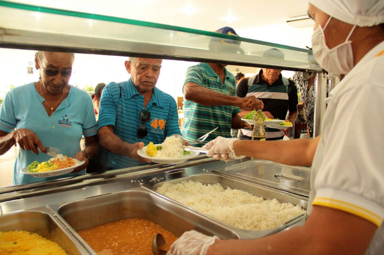 Foto mostra pessoas, incluindo idosos, se servindo em uma fila de balcão self-service em um restaurante popular.