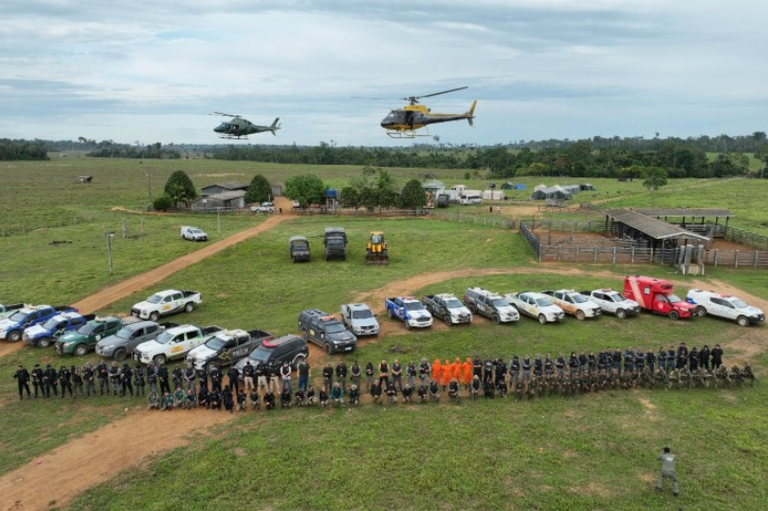 Vista aérea de uma operação de desintrusão em uma área desmatada com mata ao fundo. Em primeiro plano, uma grande formação de agentes de segurança e fiscais está alinhada em frente a uma fila de veículos (caminhonetes, SUVs) e equipamentos pesados (tratores). Dois helicópteros estão voando baixo sobre a cena. Ao fundo, estruturas rurais e cercados de gado são visíveis.