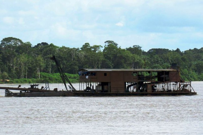 Fotografia de draga em um rio. A draga é uma embarcação usada para sugar sedimentos do leito do rio para procurar ouro. Os garimpeiros jogam mercúrio (que é tóxico) no material filtrado para separar o ouro da areia e depois descartam o metal tóxico diretamente na água.